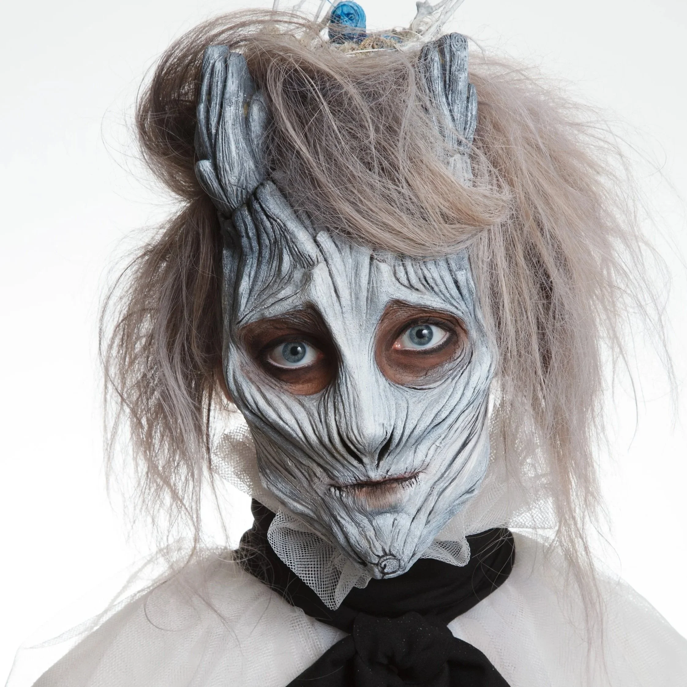 Person wearing a white and black costume with a skull-like foam latex makeup prosthetic, disheveled blonde hair, and a black necktie.