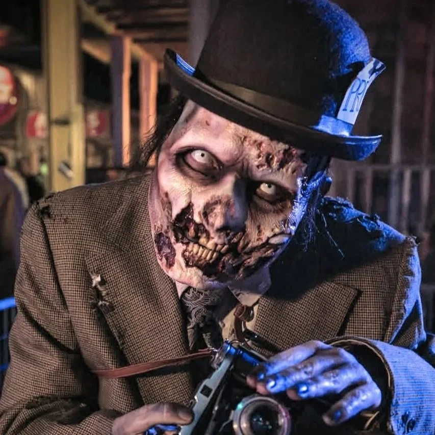 A person wearing a zombie makeup prosthetic dressed as a zombie with decayed skin and glowing eyes, wearing a vintage brown jacket and a black top hat, holding a camera.