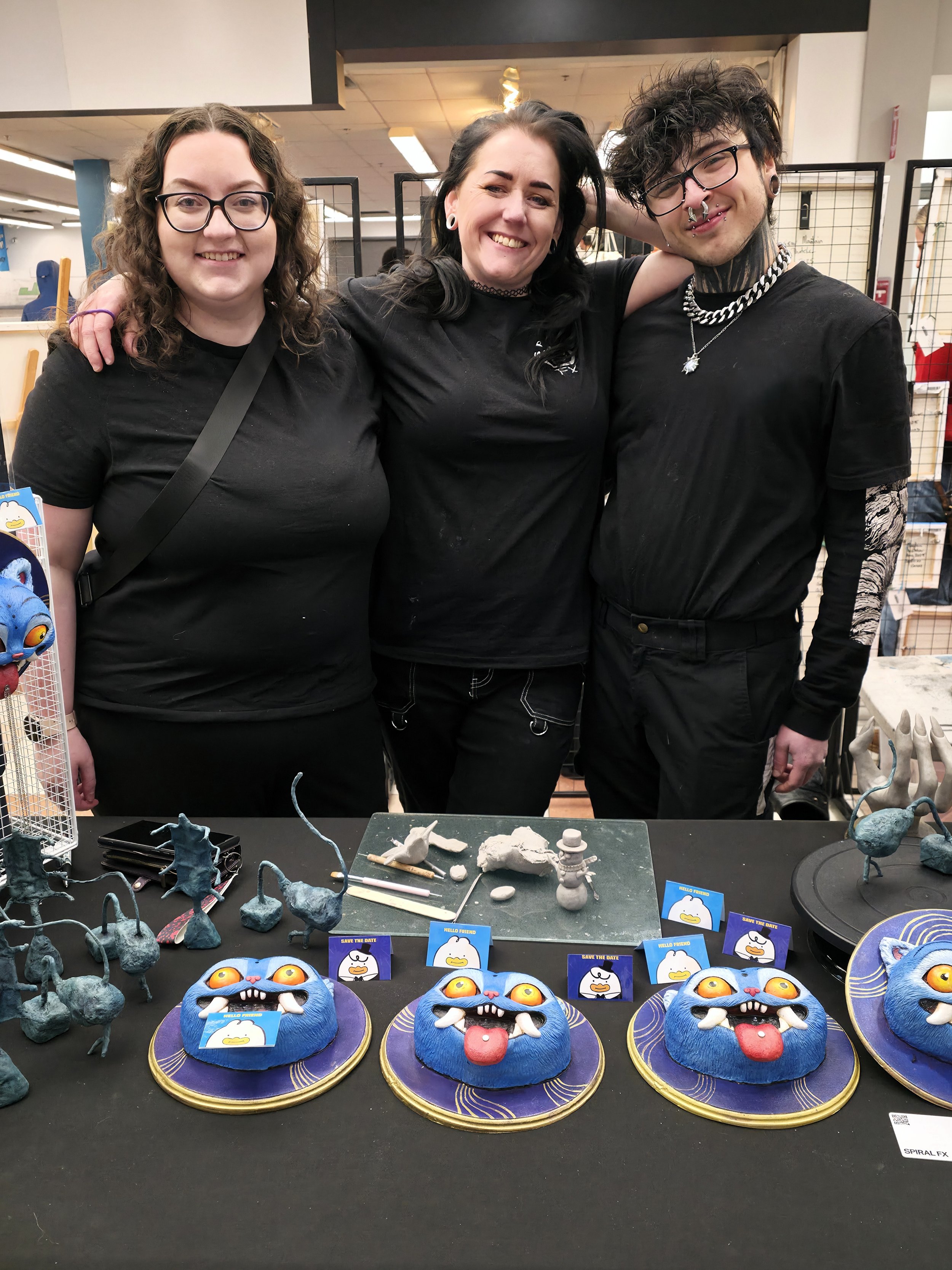 Three smiling people stand behind a table of colorful monster-themed pastries and clay sculptures at an event or expo.