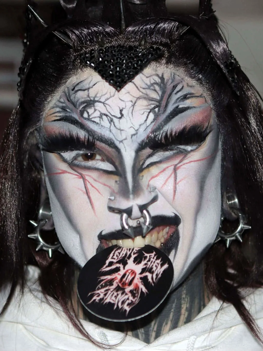 Person with elaborate horror-themed face makeup resembling an edgy drag queen, with dark hair, piercings, and a black circle sticker with a logo in their mouth.