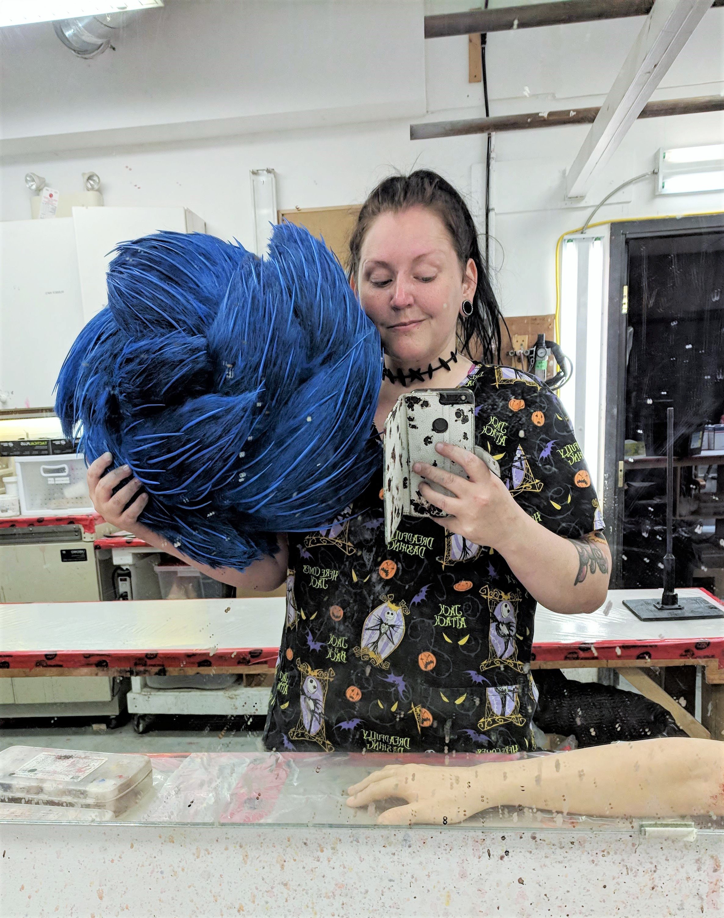 A woman with black hair, wearing a Halloween themed shirt and choker, is taking a selfie in a workshop mirror while holding a large, bright blue wig. The workshop has industrial and crafting equipment visible in the background.