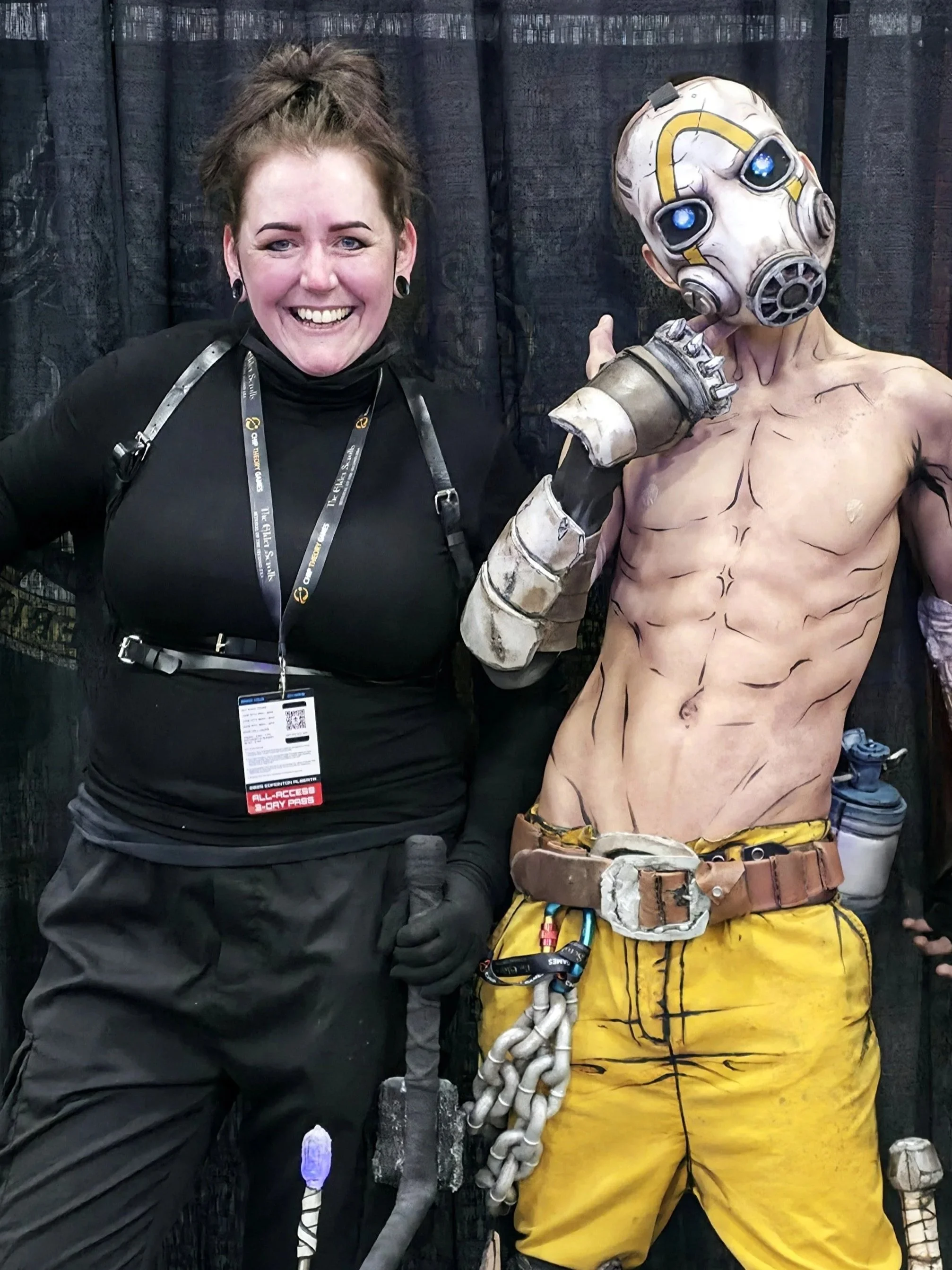 A woman smiling and standing next to a cosplayer resembling psycho bandit from borderlands with an elaborate body makeup giving the illusion of cell shading