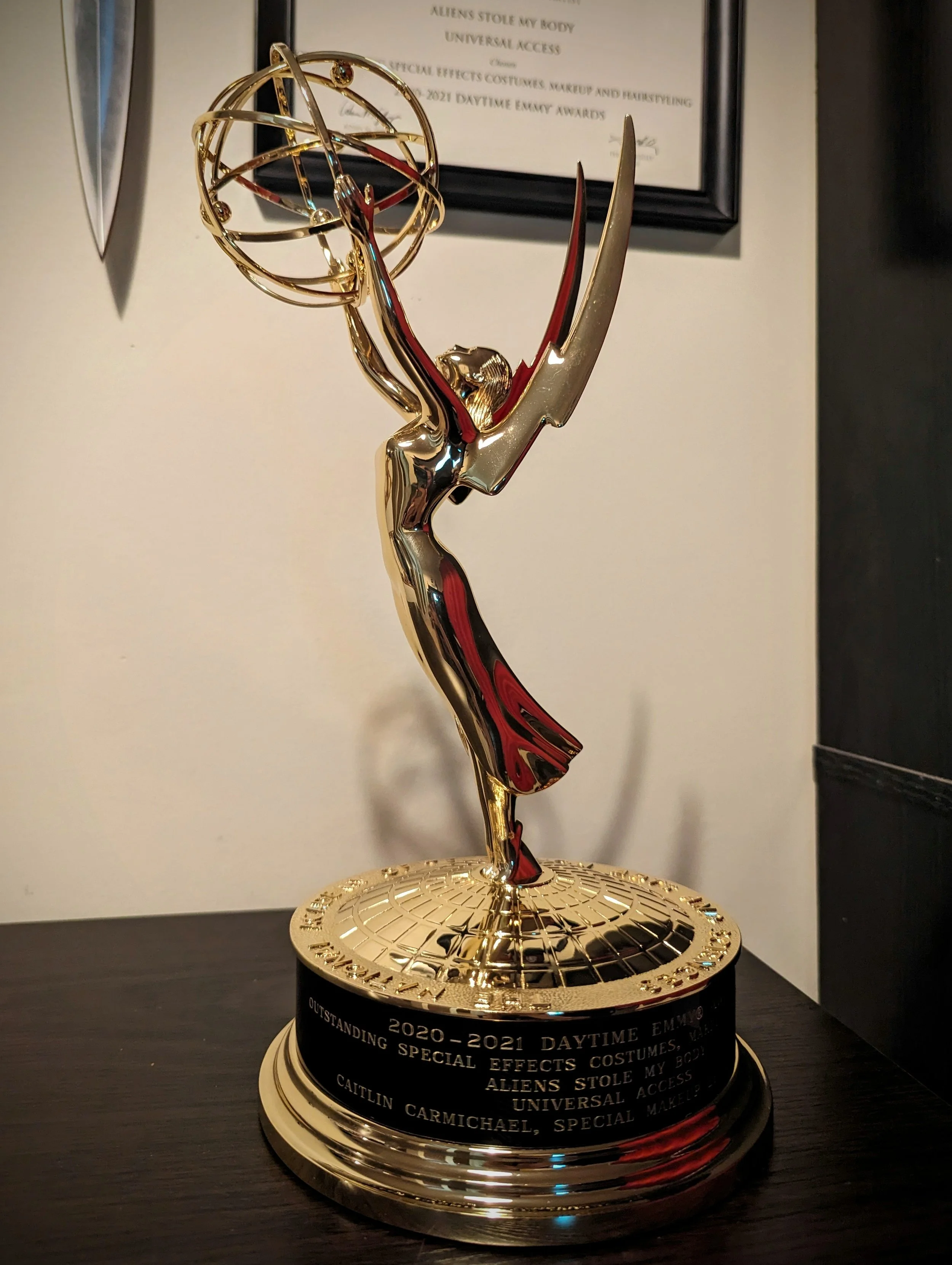 A gold Emmy award trophy with wings, a statue of a woman holding an atom at the top, on a black base with an engraved plaque.