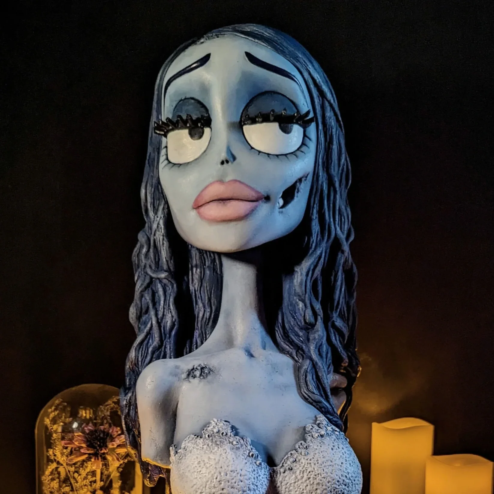 A sculpture of a stylized woman resembling the corpse bride from tim burtons movie with exaggerated facial features, long wavy hair, large lips, and prominent eyes, set against a dark background with some decorative items and yellow candles nearby.