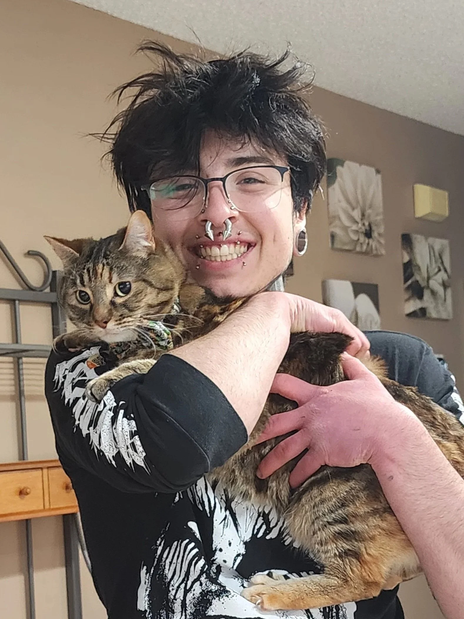 A person with dyed black hair, glasses, and multiple facial piercings holding a tabby cat.