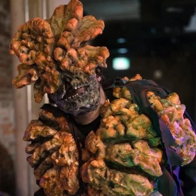Person wearing a costume with large, irregularly shaped orange and green organ-like growths covering their head, face, and shoulders, resembling a clicker from the last of us