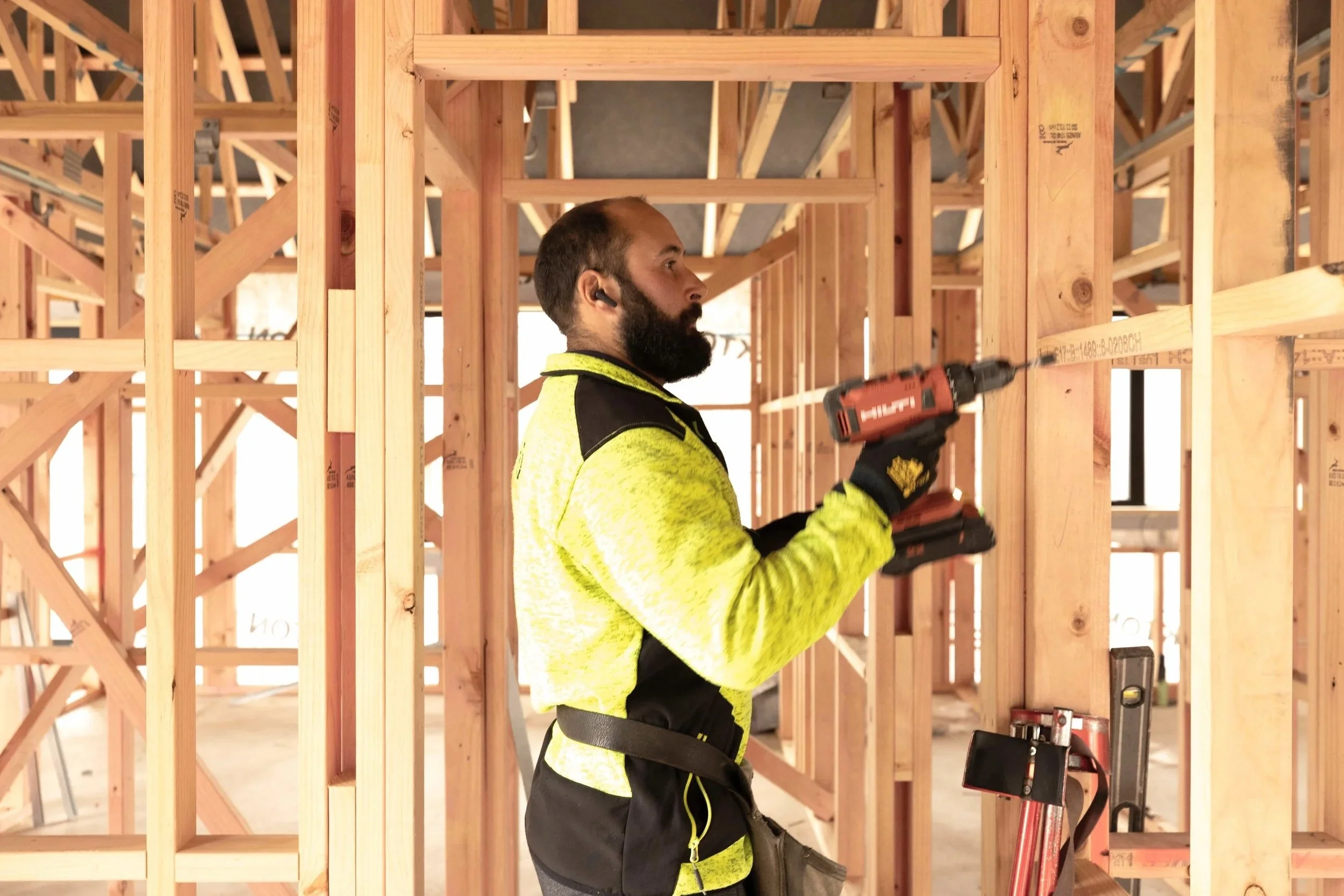 A man with a beard and short hair wearing a yellow high-visibility jacket, black gloves, and ear protection, using a cordless drill on a wooden framing at a construction site.