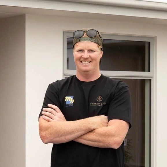 A smiling man standing outdoors in front of a house, wearing a black t-shirt, sunglasses on a cap, and crossing his arms.