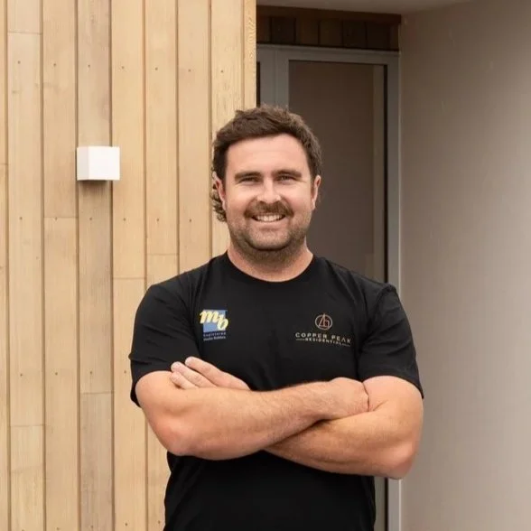 Man with dark curly hair and beard smiling with arms crossed, wearing a black t-shirt, standing indoors against a wood-paneled wall.
