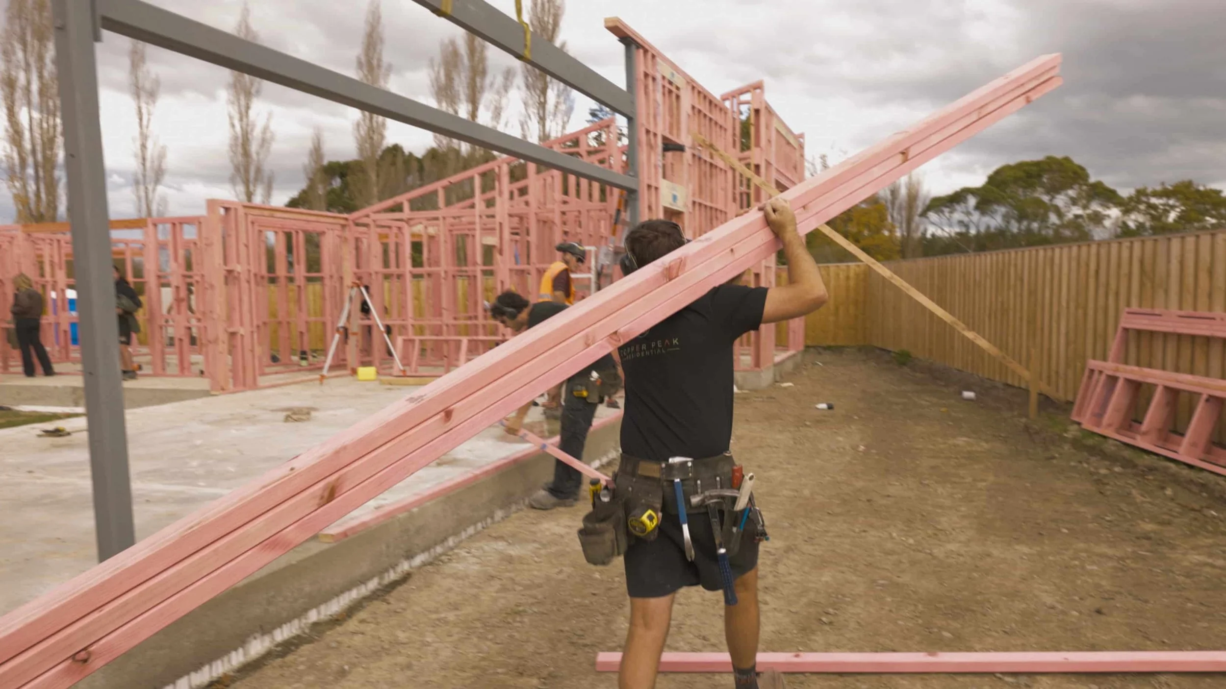 Construction workers building a house frame with pink wood. A man in the foreground is carrying a long pink wooden beam, wearing a tool belt and shorts. Other workers are visible in the background working on the structure, which is partially built with framing and roofing in progress. The site is outdoors on a cloudy day with trees and a wooden fence in the background.