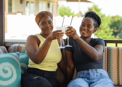 Two women are sitting on a porch, enjoying drinks together and smiling at the camera.