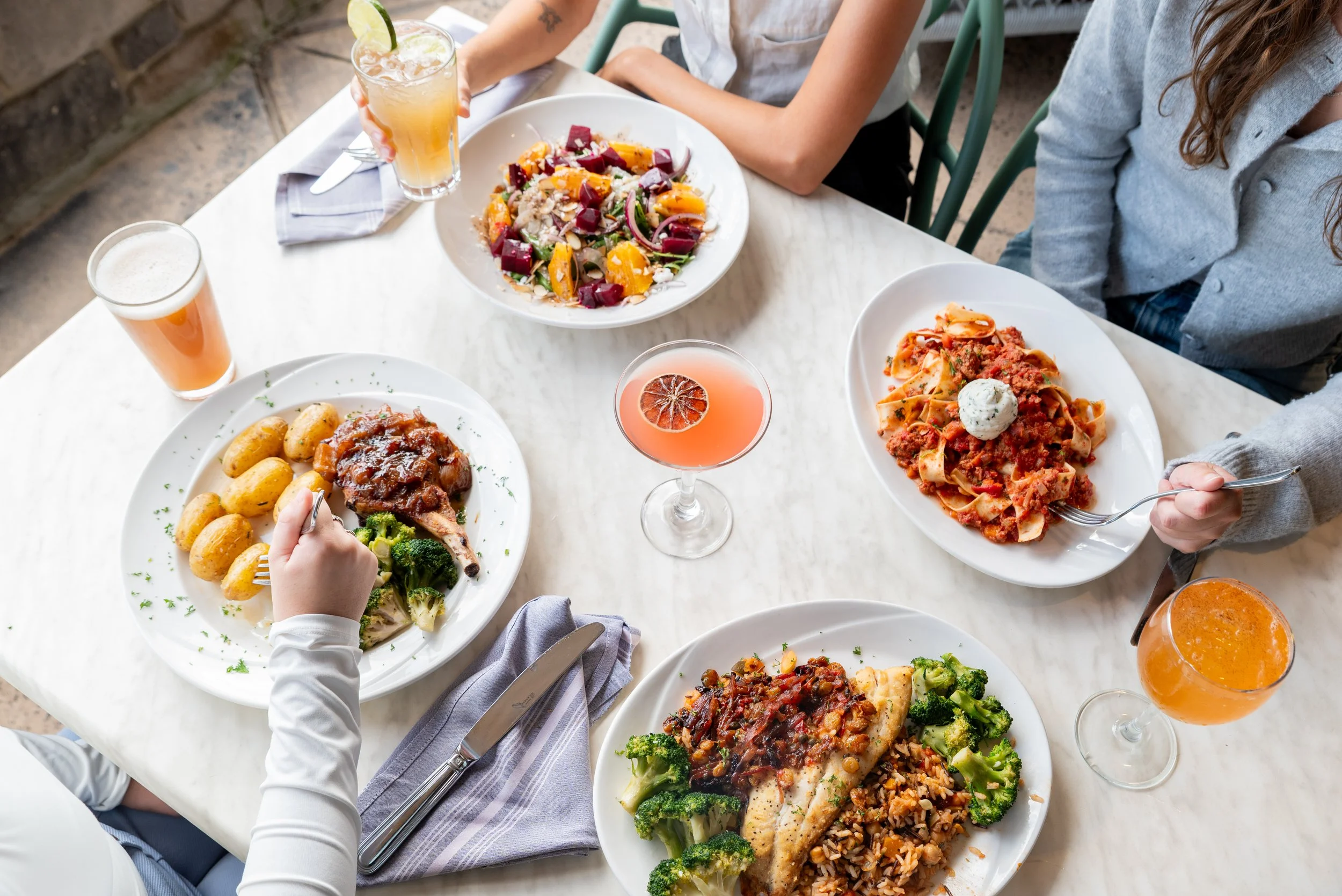 A restaurant table with multiple plates of food, drinks, and some people dining, including a bowl of vegetable salad, a plate with chicken and potatoes, a pasta dish with meat sauce, and a fish fillet with broccoli and rice, along with cocktails and beer.