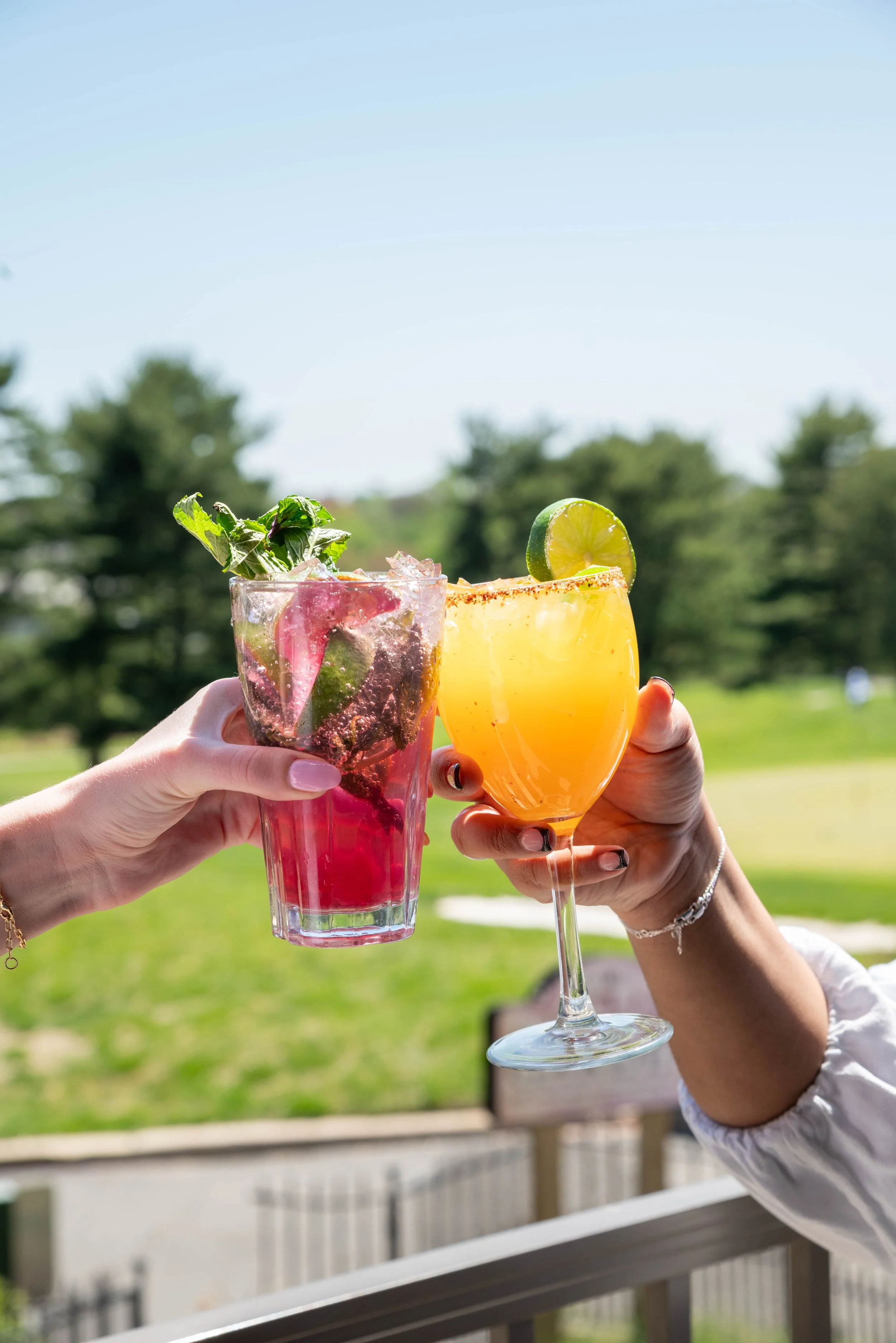 Two people clinking glasses of colorful cocktails outdoors on a sunny day, with a grassy field and trees in the background.
