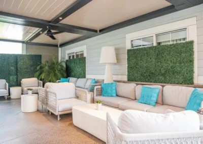 Outdoor patio with beige couches and chairs, blue pillows, white side table, white lamp, potted plants, and green hedge panels on the wall.