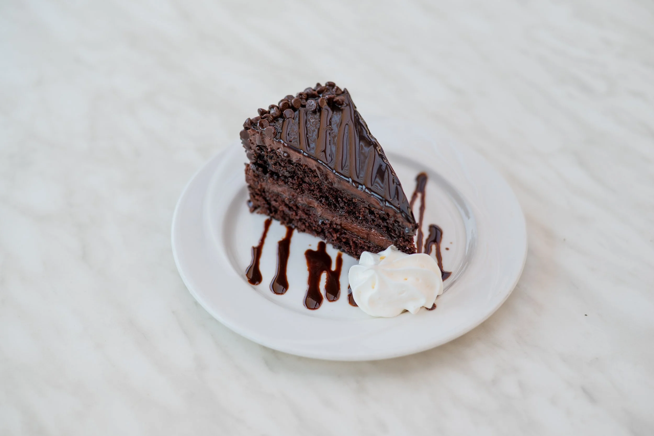 Slice of chocolate cake with glazed frosting and chocolate chips, served with whipped cream on a white plate.