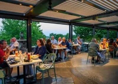 People dining at outdoor restaurant with tables, chairs, and a metal roof, surrounded by green trees.