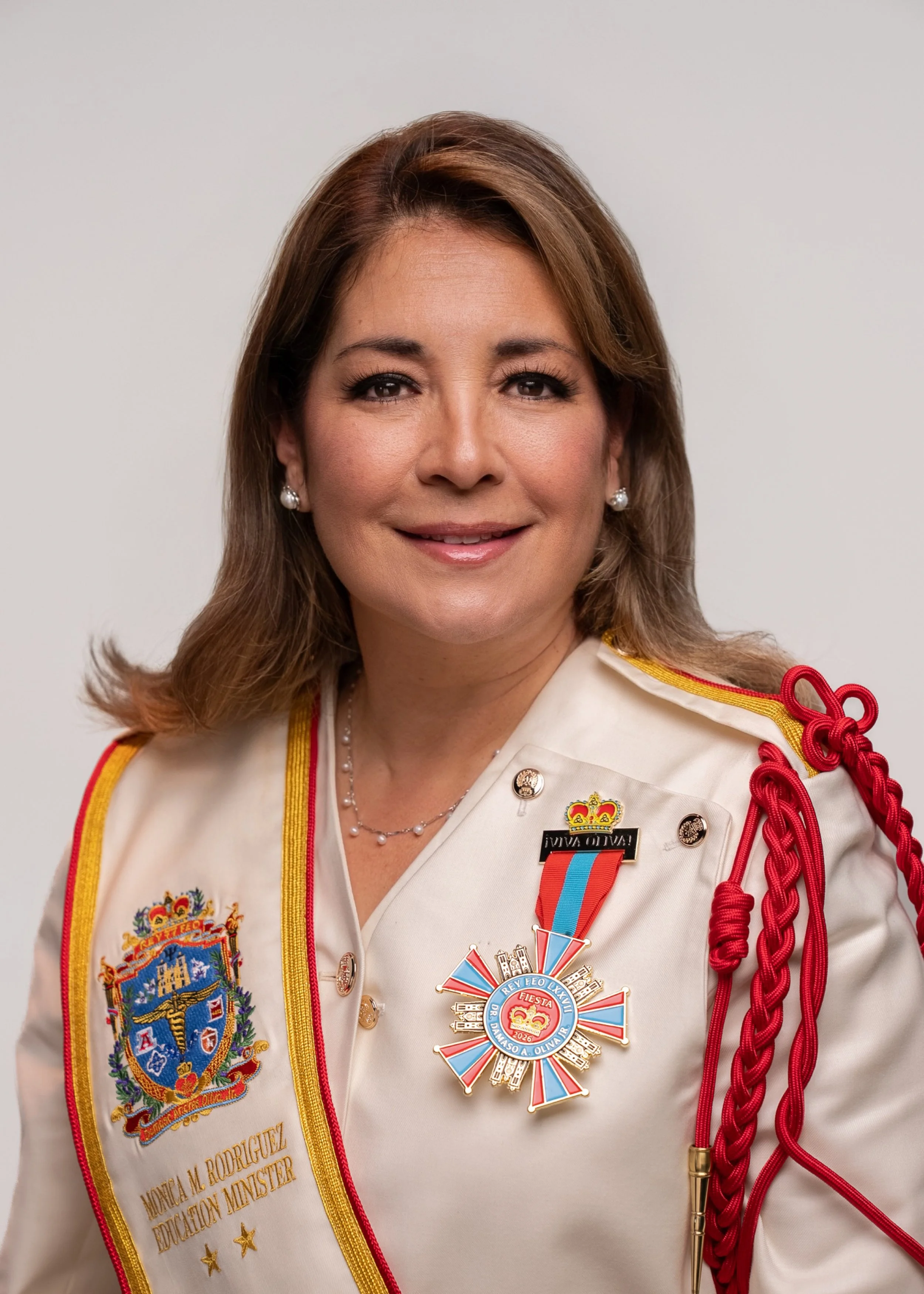 A woman with shoulder-length brown hair, styled with highlights, wearing a red button-up shirt and jewelry, smiling at the camera against a plain gray background.