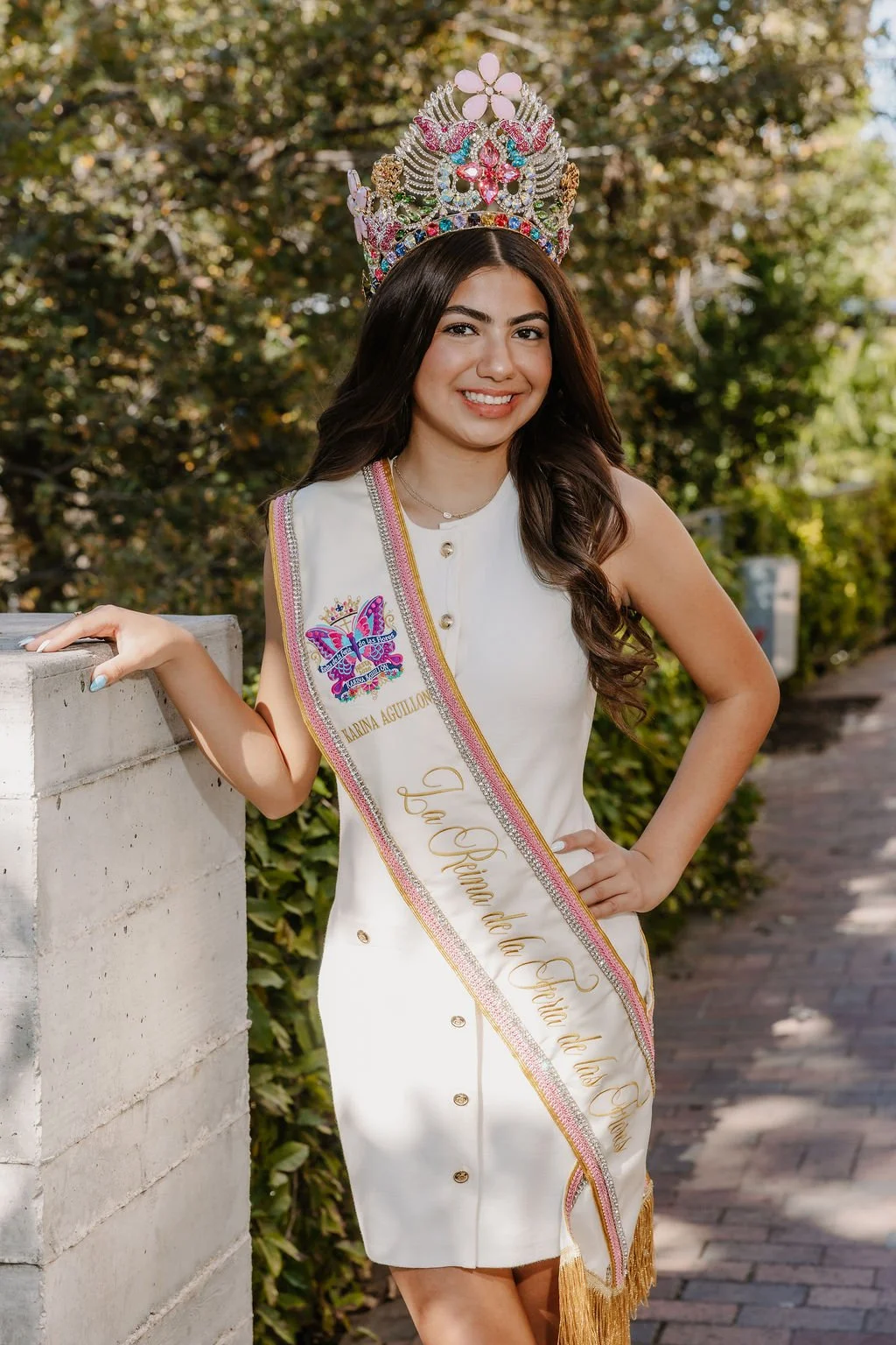 Smiling young woman with long dark hair, wearing a light blue off-shoulder dress with a floral pattern and a gold cross necklace, against a plain gray background.