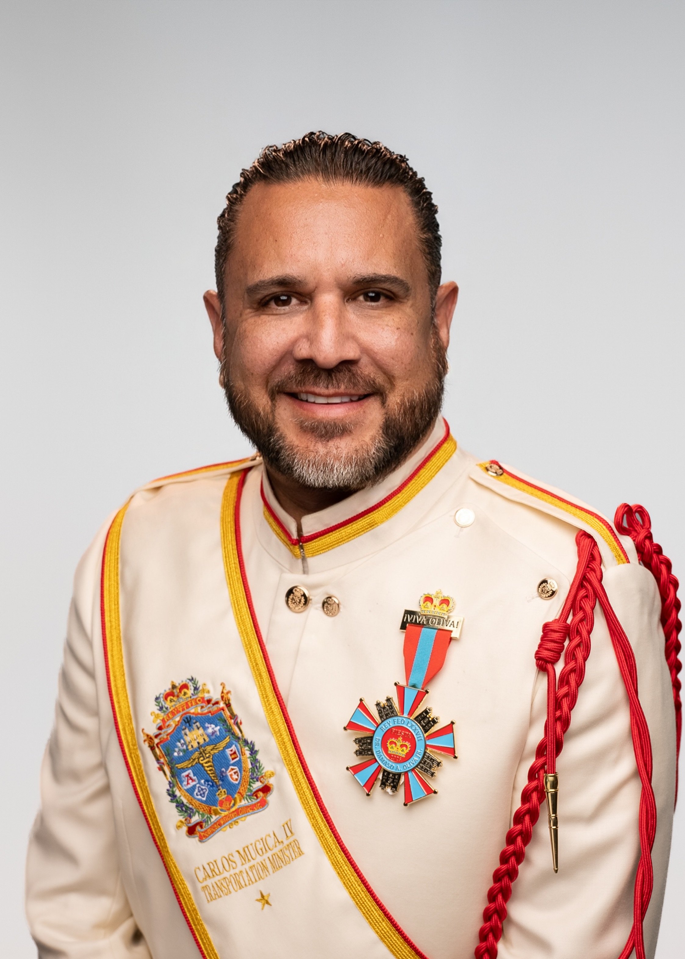 A man with short, dark hair and a beard, wearing a red shirt and a gold chain necklace, smiling at the camera against a plain light gray background.