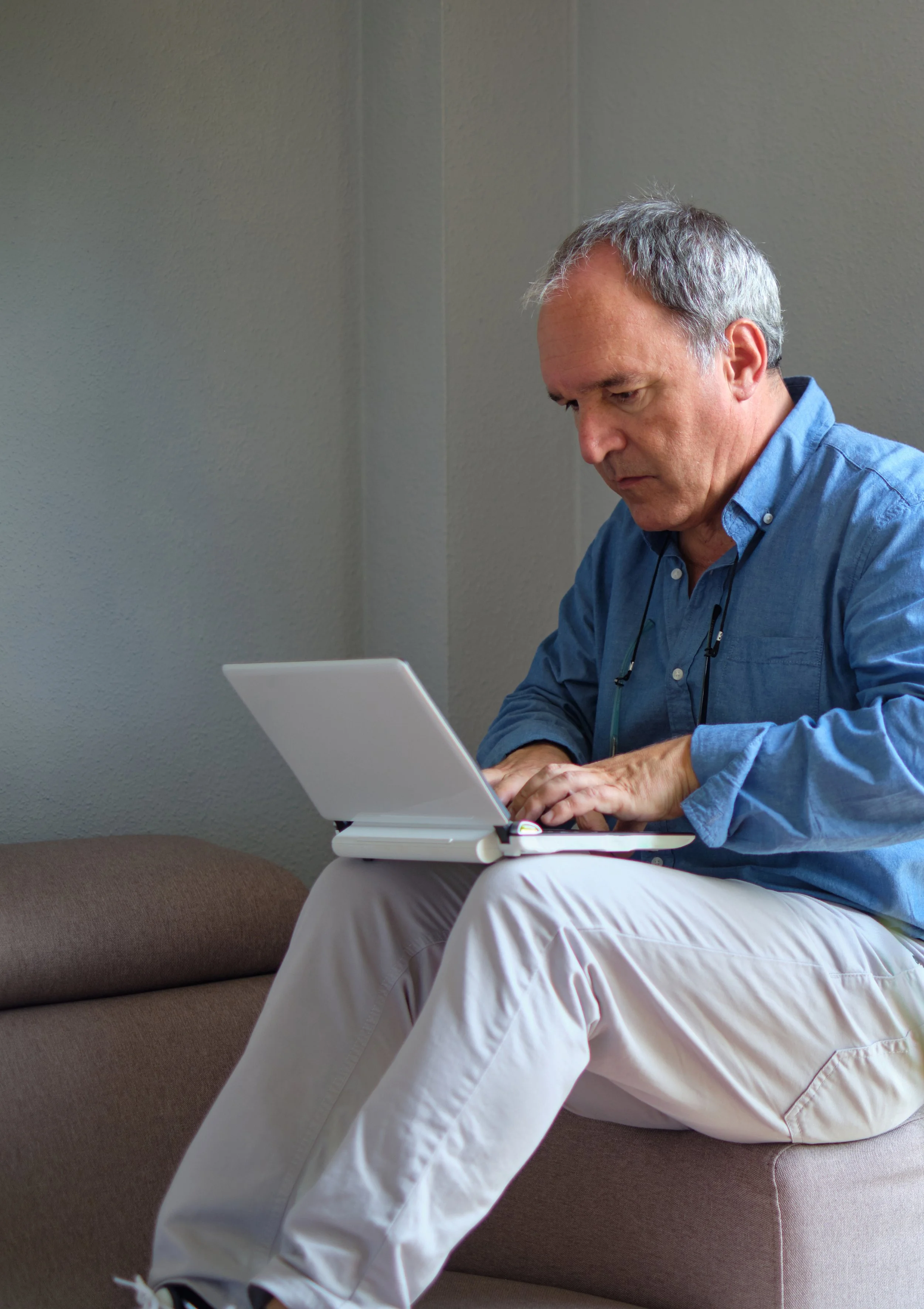 Older man with gray hair in a blue shirt sitting on a brown couch, working on a small white laptop, focused on the screen.