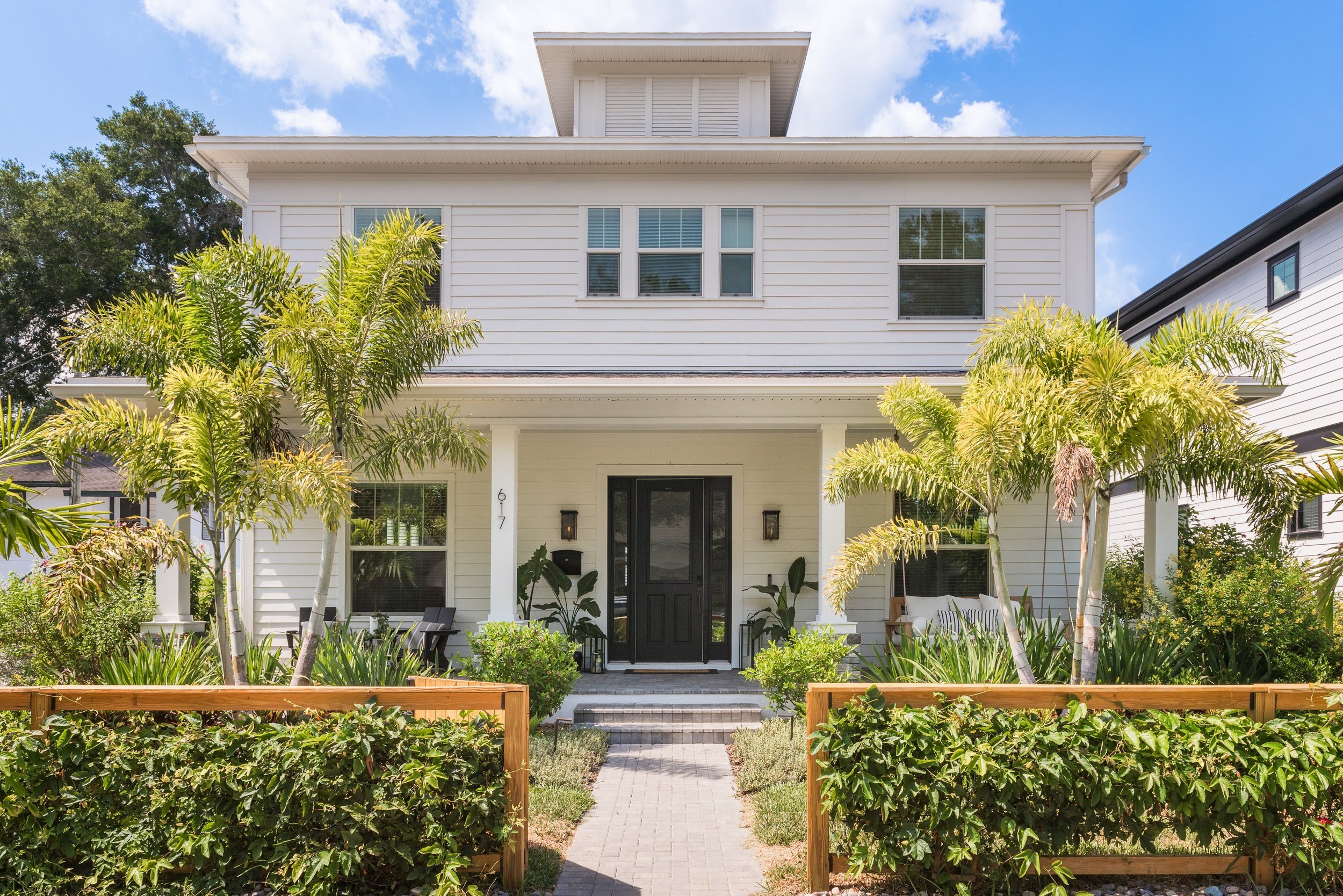 Two-story white house with black door and windows, surrounded by palm trees and green shrubs, under a blue sky.