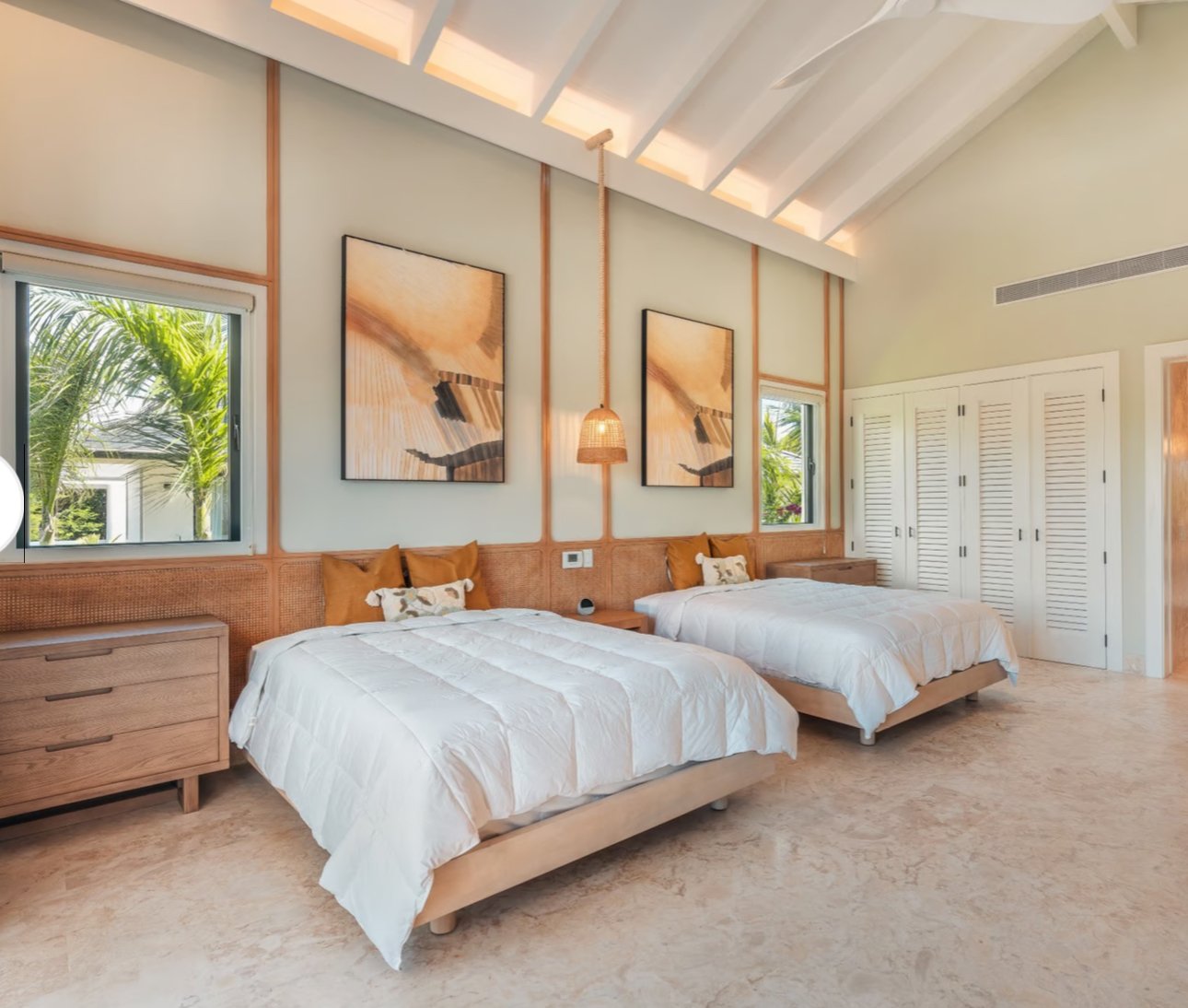 Modern bedroom with two beds, white bedspreads, and wooden furniture. Natural light floods from a window showing palm leaves outside. Walls feature abstract artwork, a hanging lamp, and a slanted ceiling with exposed beams.