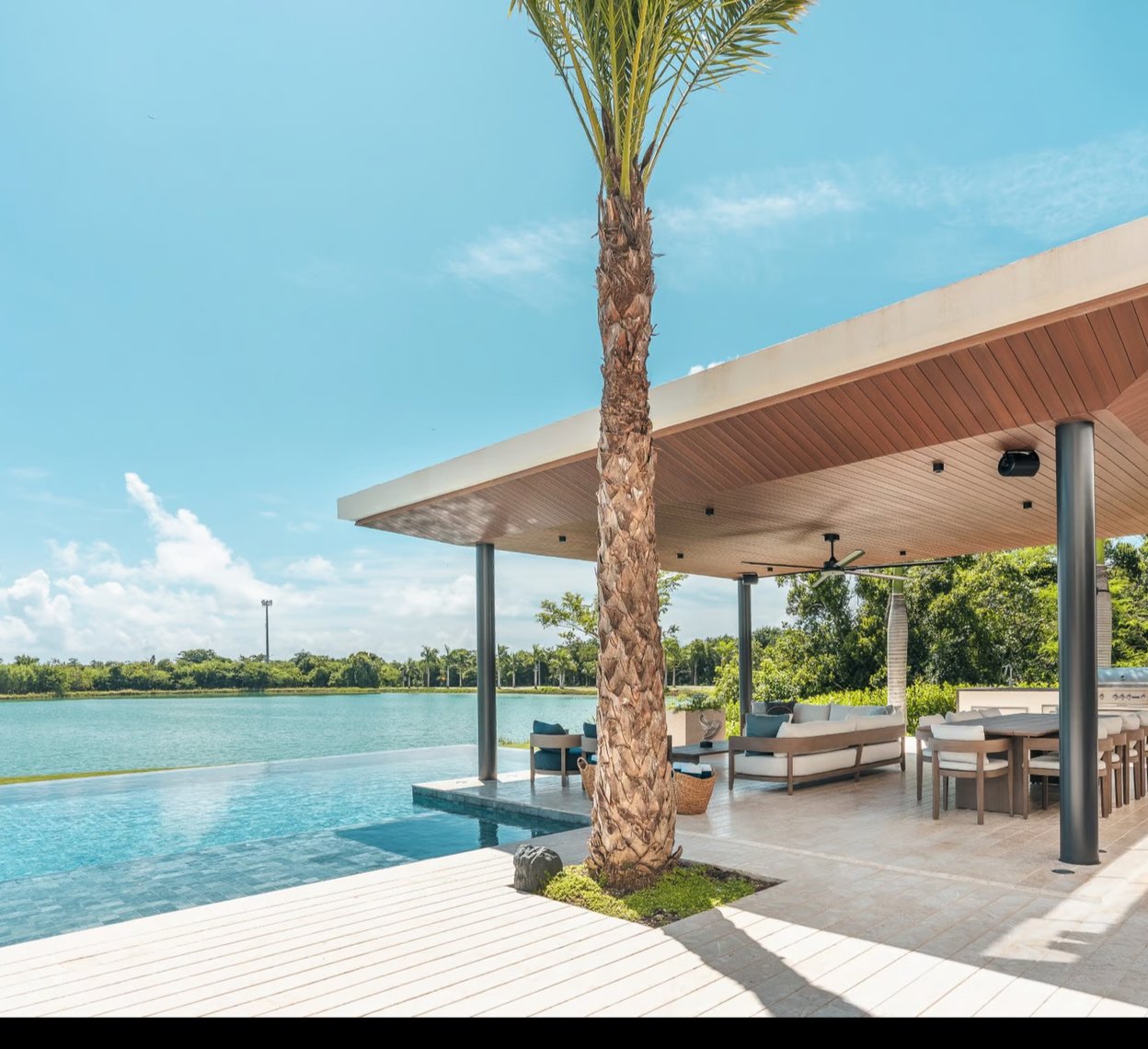 Modern patio with a pool and lake view, featuring a palm tree, wooden pergola, outdoor seating, and dining area.