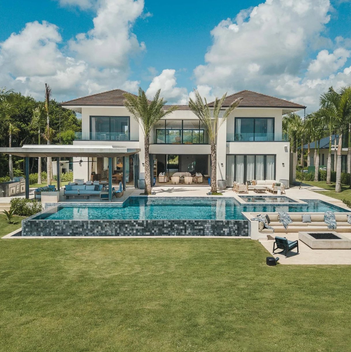 Luxury modern villa with a large infinity pool, outdoor seating area, and palm trees in a tropical setting.