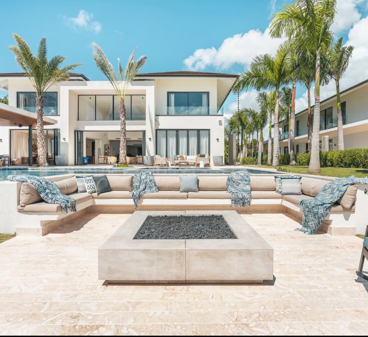 Luxury modern mansion with outdoor seating area, fire pit, swimming pool, and palm trees under a clear blue sky.