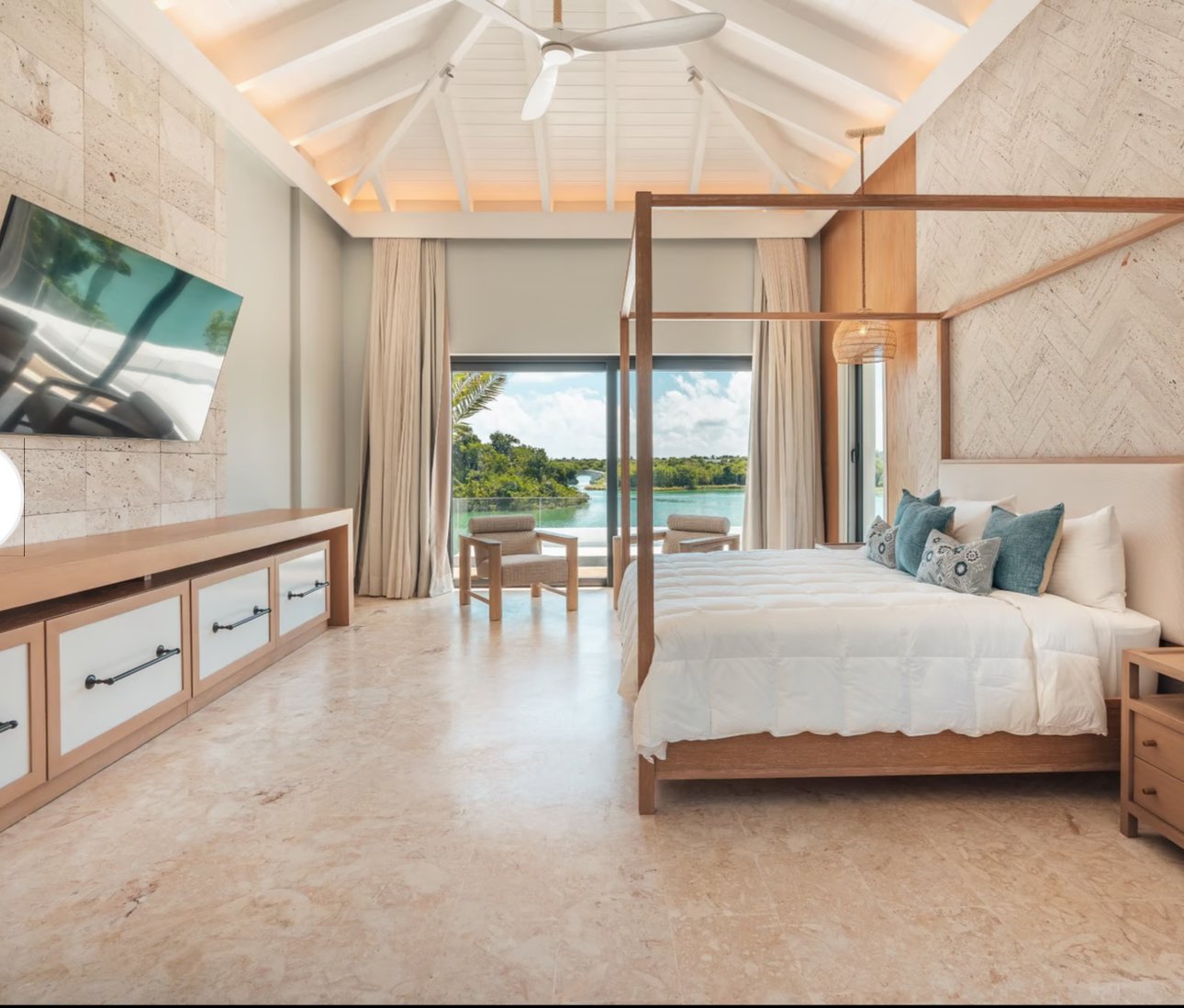 Luxurious bedroom interior with a canopy bed, light wooden furniture, and a large flat-screen TV. The room features high ceilings with fan, large windows offering a scenic water view, and light-colored tiled floors.