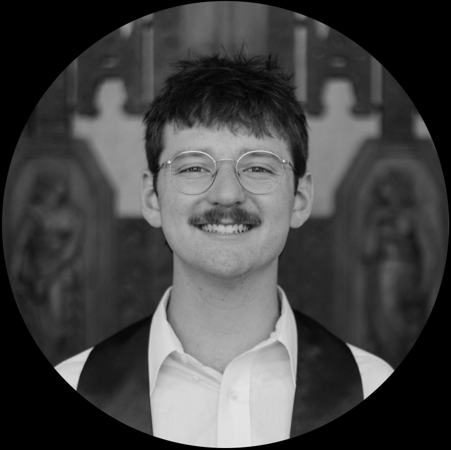 Young man with glasses, mustache, and brown hair, smiling, wearing a white shirt and maroon vest, standing in front of wooden carvings or artwork.