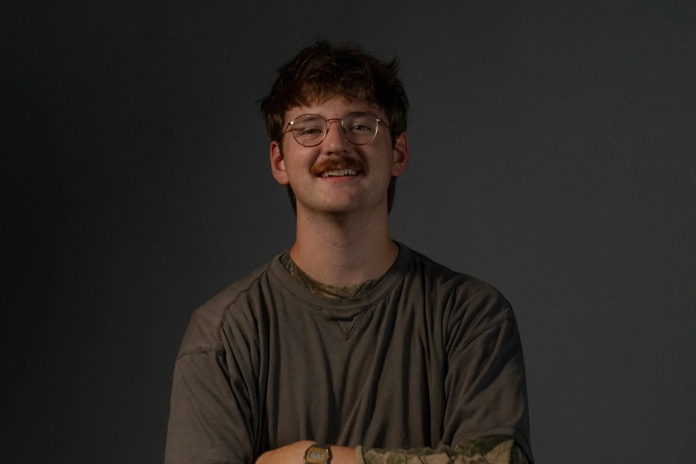 A man with glasses, a mustache, and a smile, wearing a white shirt and a dark vest, standing in front of a carved wooden background.