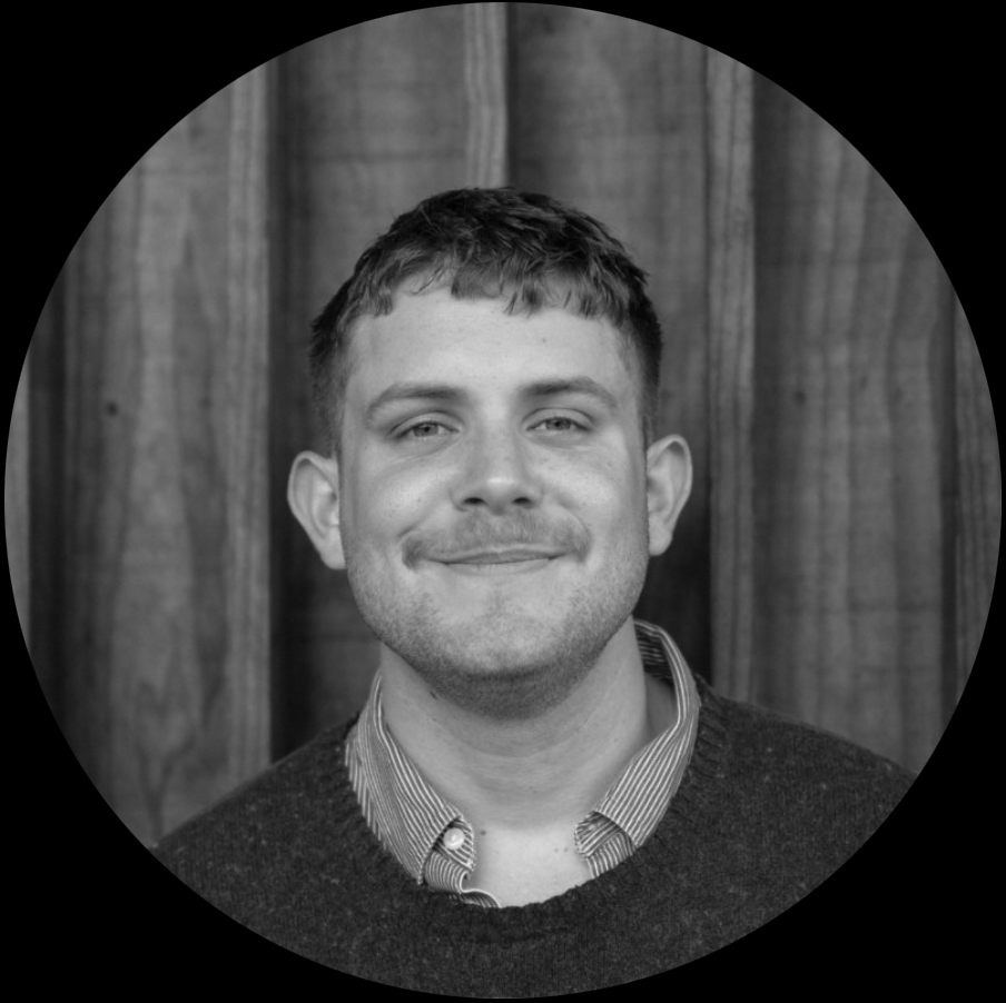 Black and white portrait of a man with short hair, smiling slightly, wearing a collared shirt and sweater in front of a wooden wall.