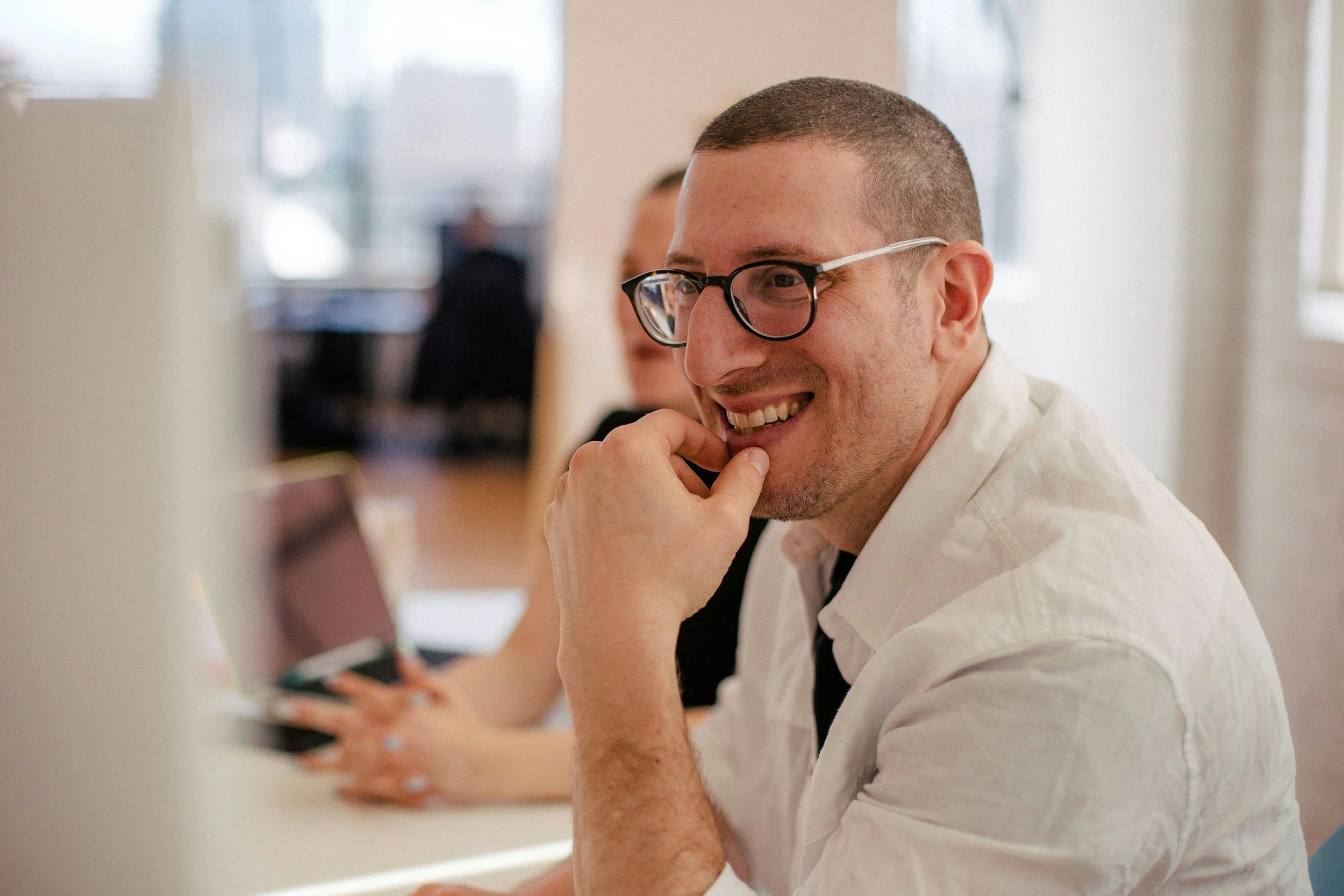 A man wearing glasses, smiling, and resting his chin on his hand, sitting at a desk with a laptop.