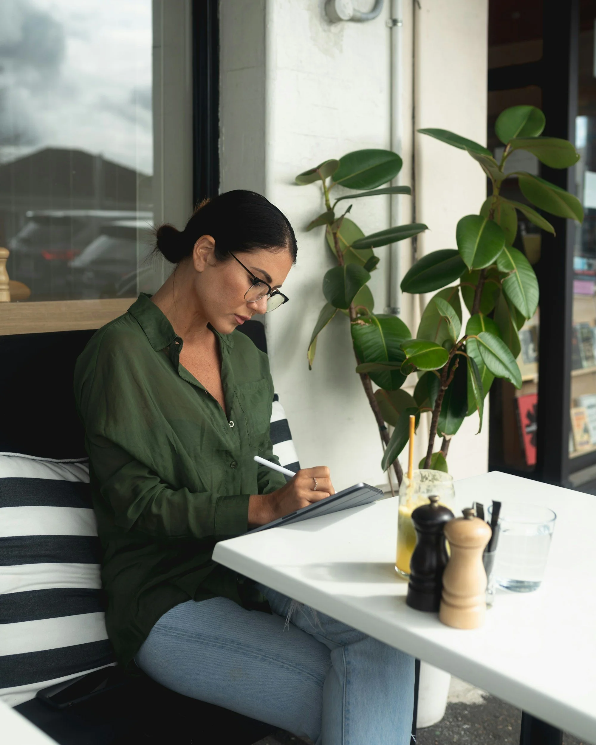 Woman with black hair and glasses in a green shirt, sitting at a table writing in a notebook with a drink with a straw