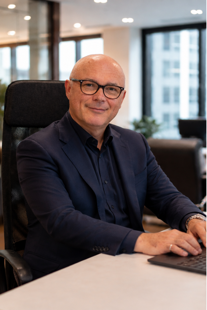 A bald man with glasses wearing a dark suit and black shirt sitting at a desk in a modern office with large windows.