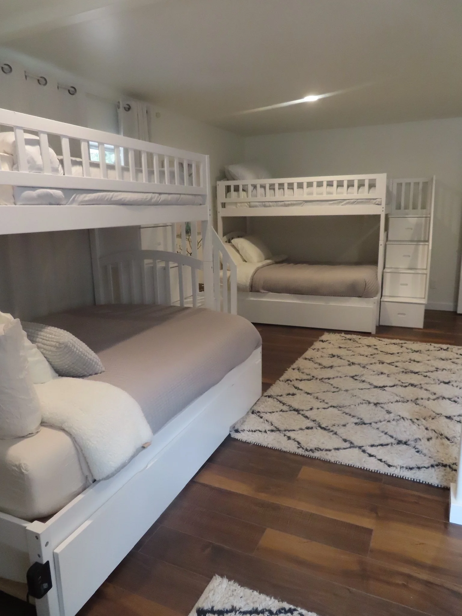 Room with two sets of white bunk beds on hardwood floor, patterned area rug, and a window with curtains.