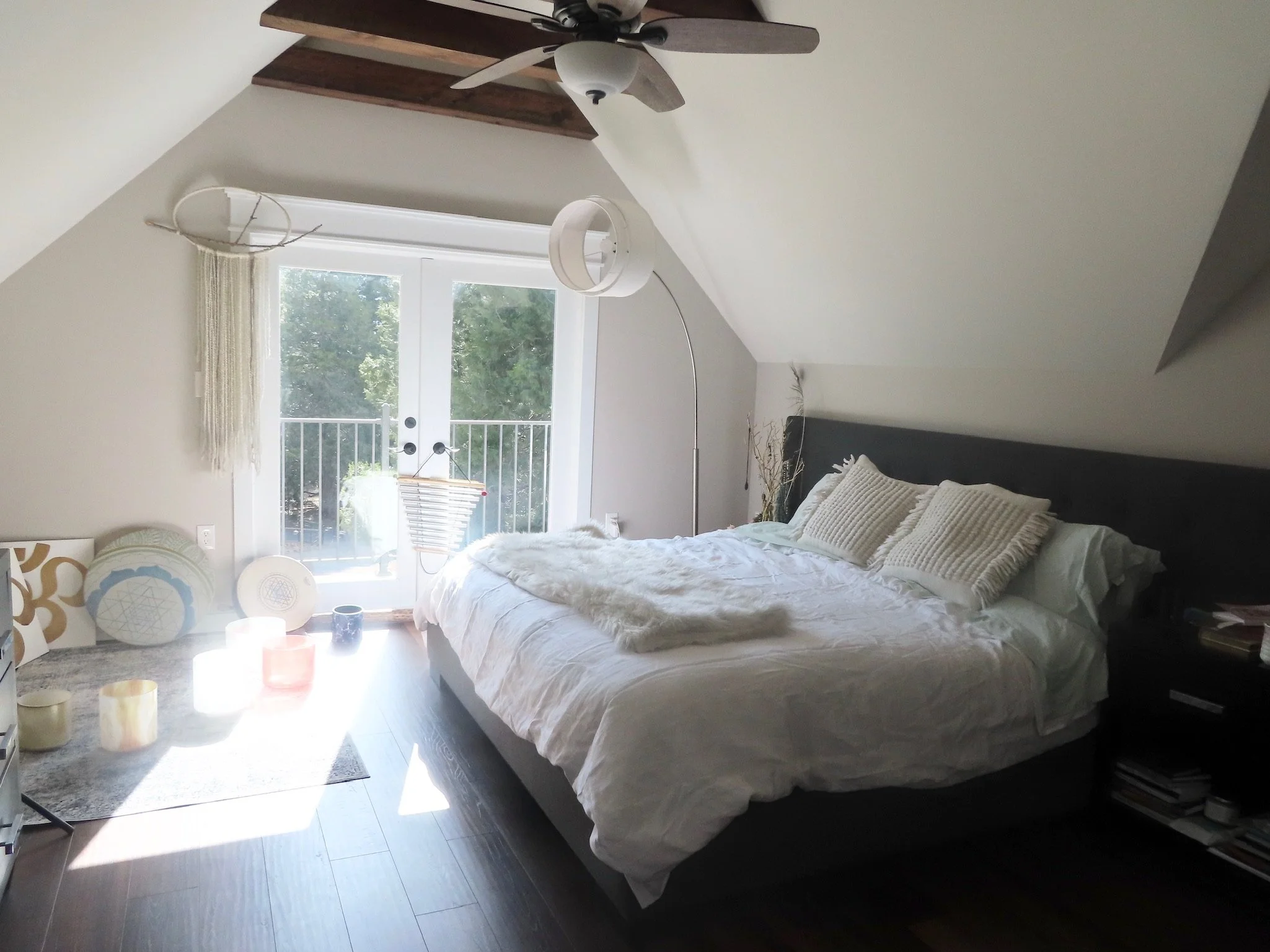 Cozy bedroom with sloped ceiling and large bed, featuring white bedding and decorative pillows, a ceiling fan, a standing lamp, and double glass doors leading to a balcony with greenery visible outside.
