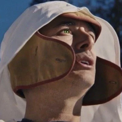 Person wearing a hood with a hollowed-out face mask or goggles, looking upward.