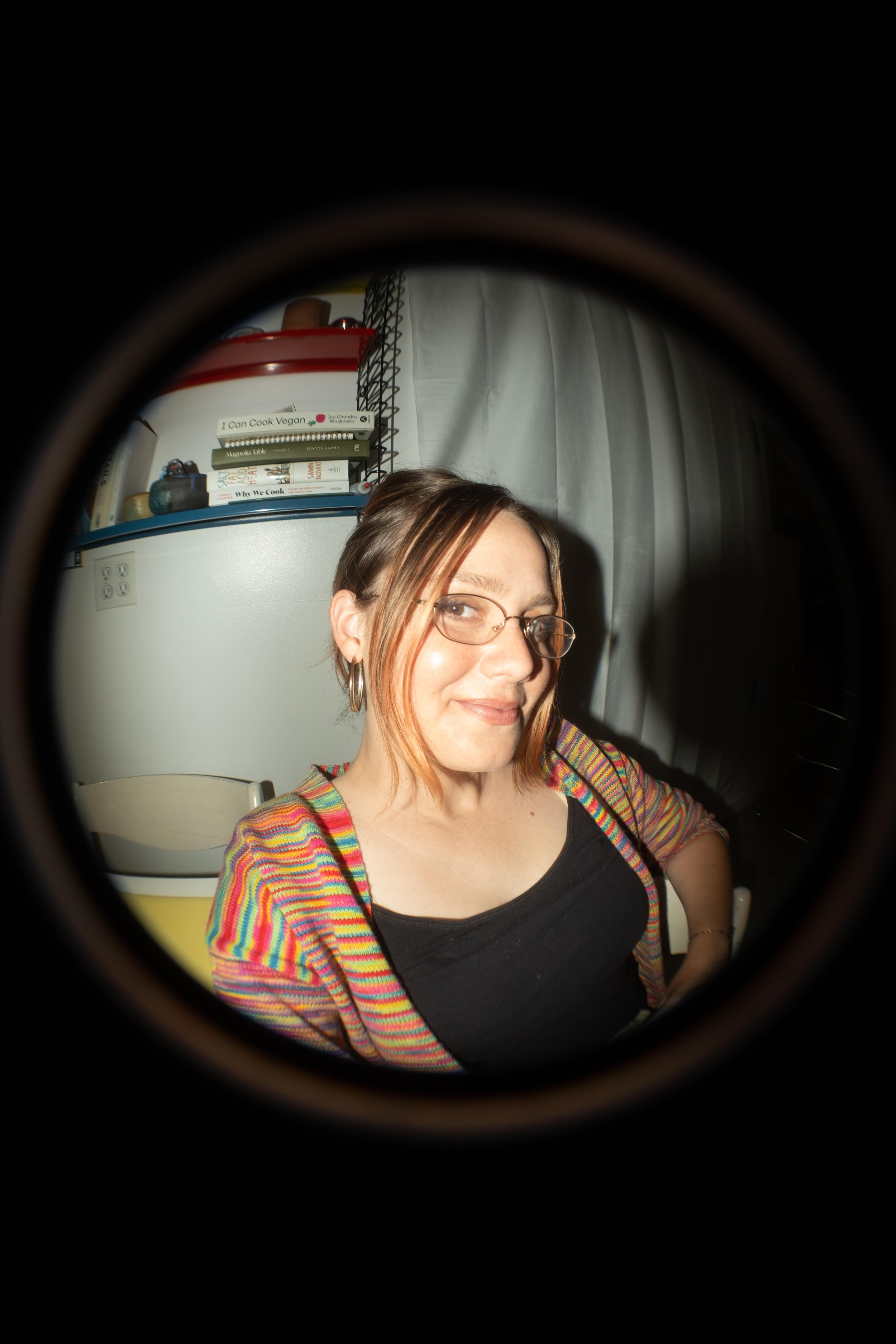 A woman wearing glasses and a colorful striped sweater sitting in a room, seen through a circular peephole.