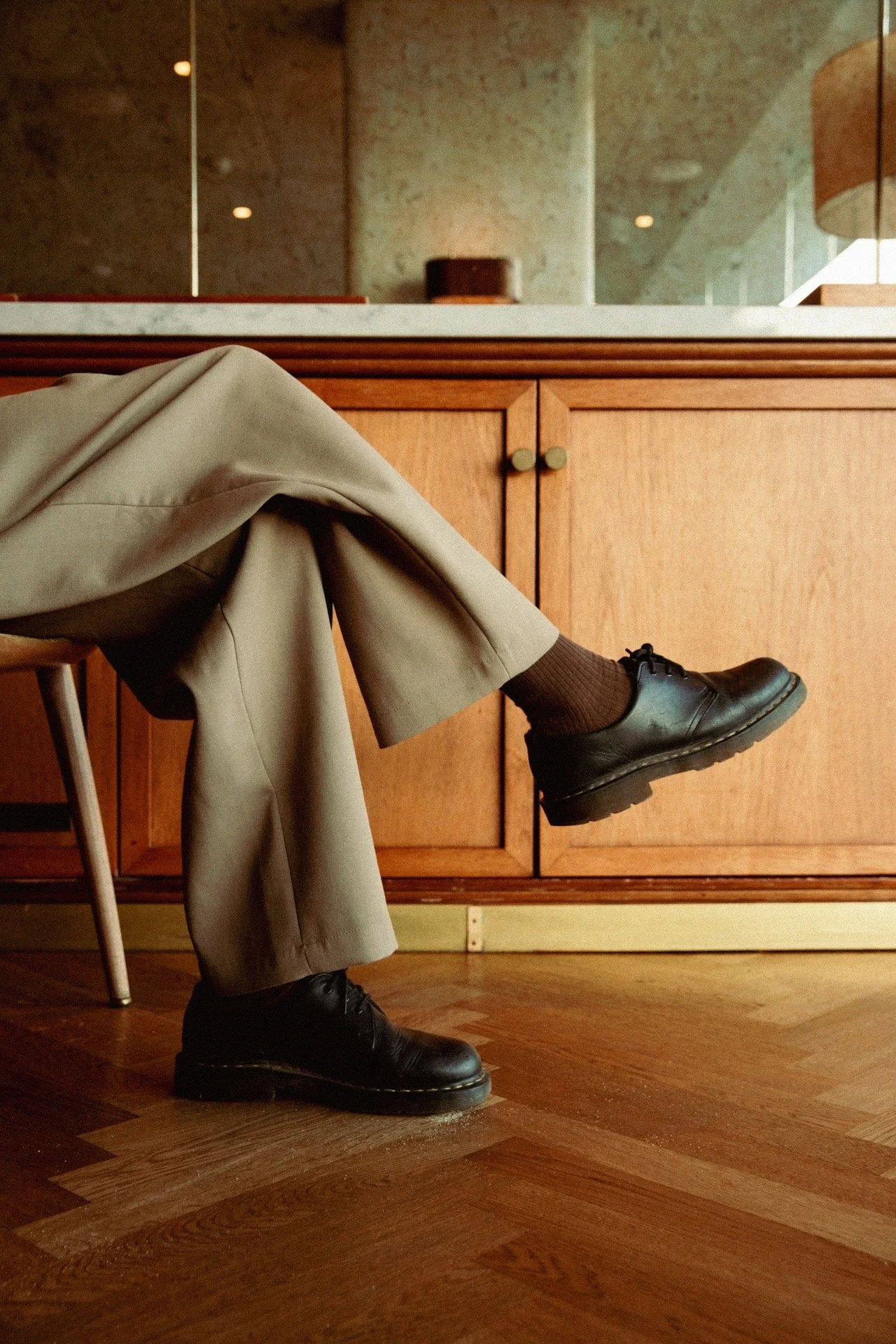 Person's legs and feet visible, seated in a room with wooden flooring and cabinetry, wearing beige pants, brown socks, and black shoes, with a mirror and a window in the background.