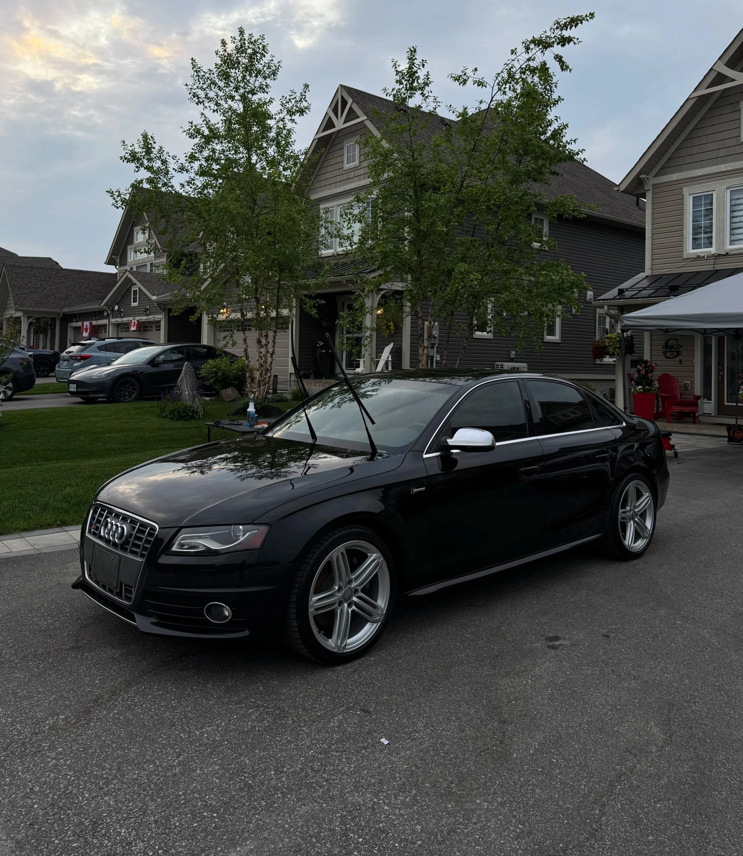 A black Audi sedan parked on a residential street in front of houses with trees and other cars visible in the background.