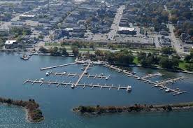 Aerial view of a marina with several docks extending into a body of water, with a city and greenery in the background.