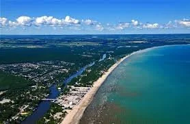 Aerial view of a coastal city with a long sandy beach, ocean, and river running parallel to the shoreline under a clear blue sky.
