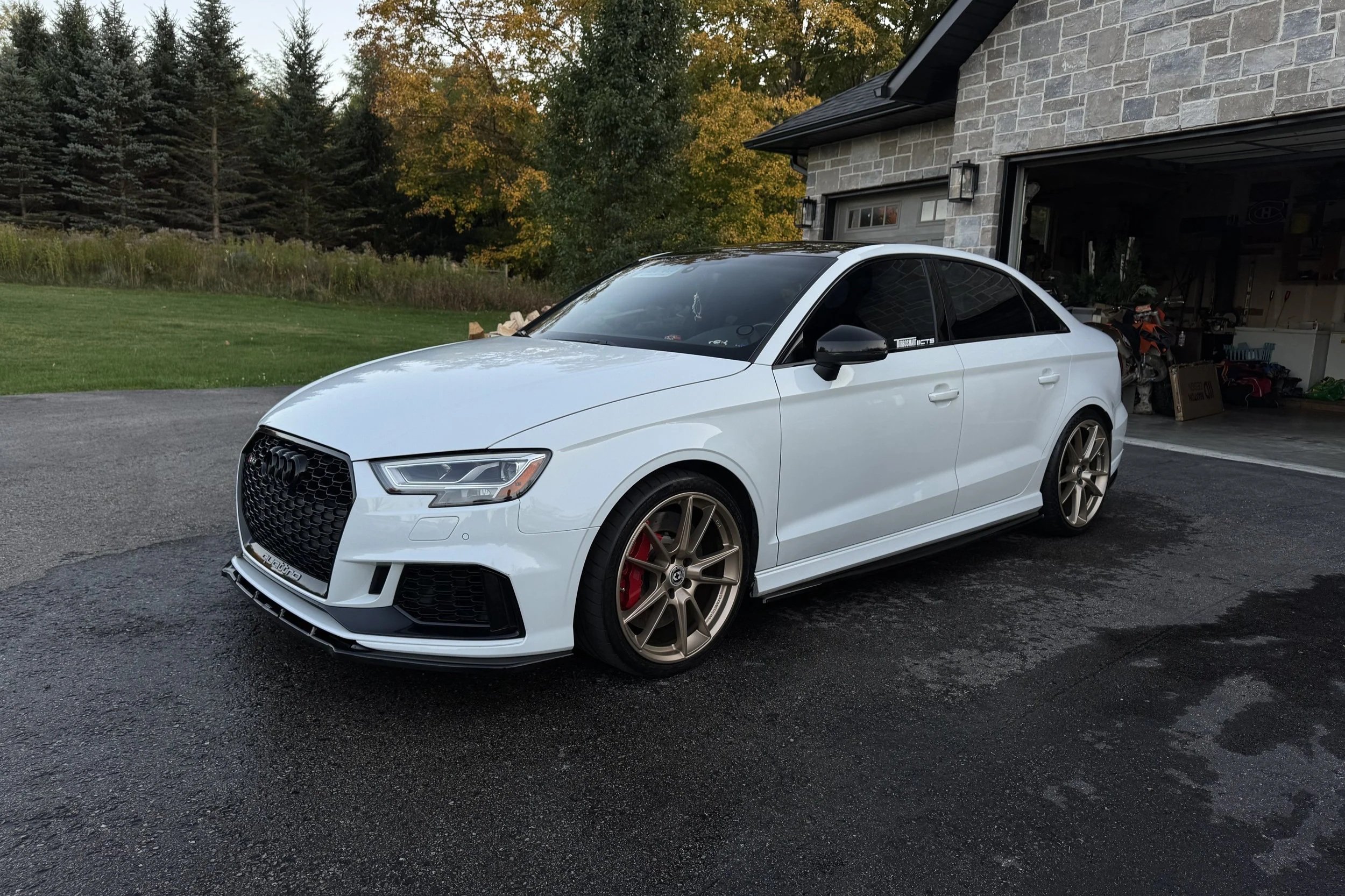 A white Audi sedan with black tinted windows and bronze alloy wheels parked on a driveway in front of a house with a stone exterior, with trees and grass in the background.