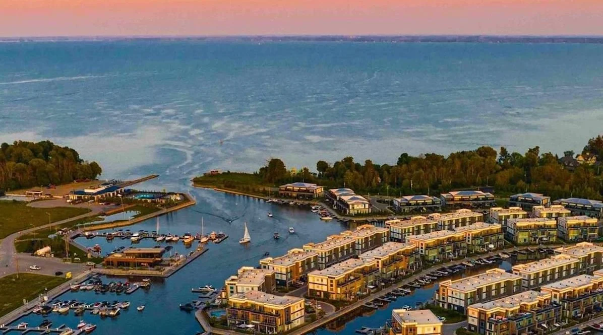 Aerial view of a marina with boats docked, residential buildings, and a large body of water in the background during sunset.