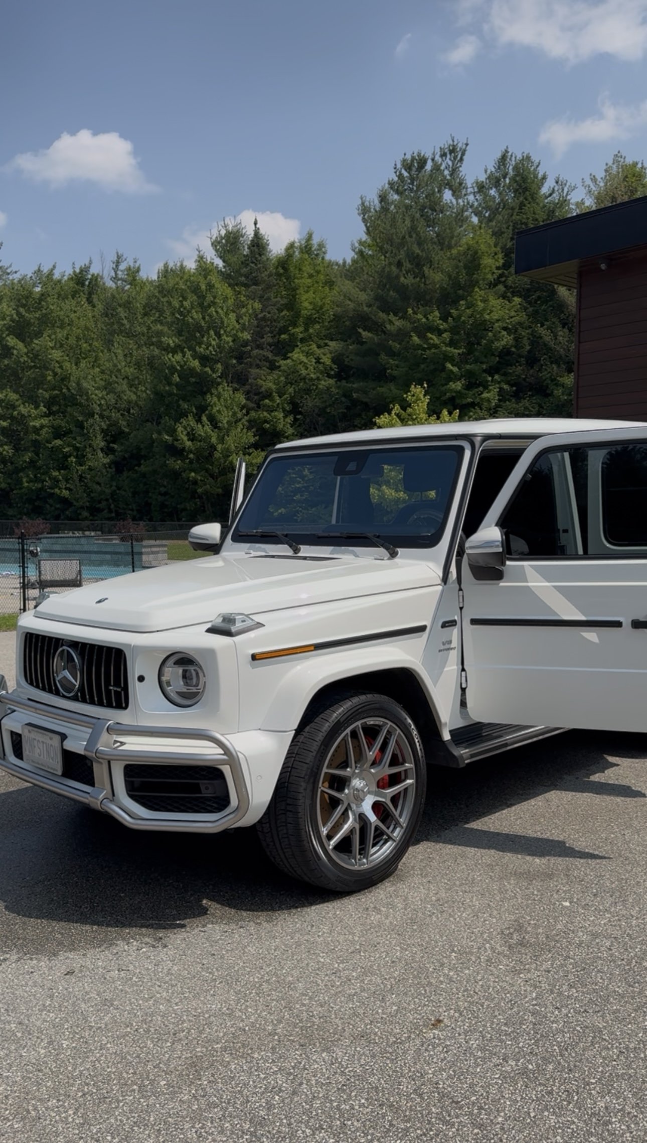A white Mercedes-Benz G-Class SUV parked on a paved surface with a background of trees, a chain-link fence, and a blue sky with clouds.