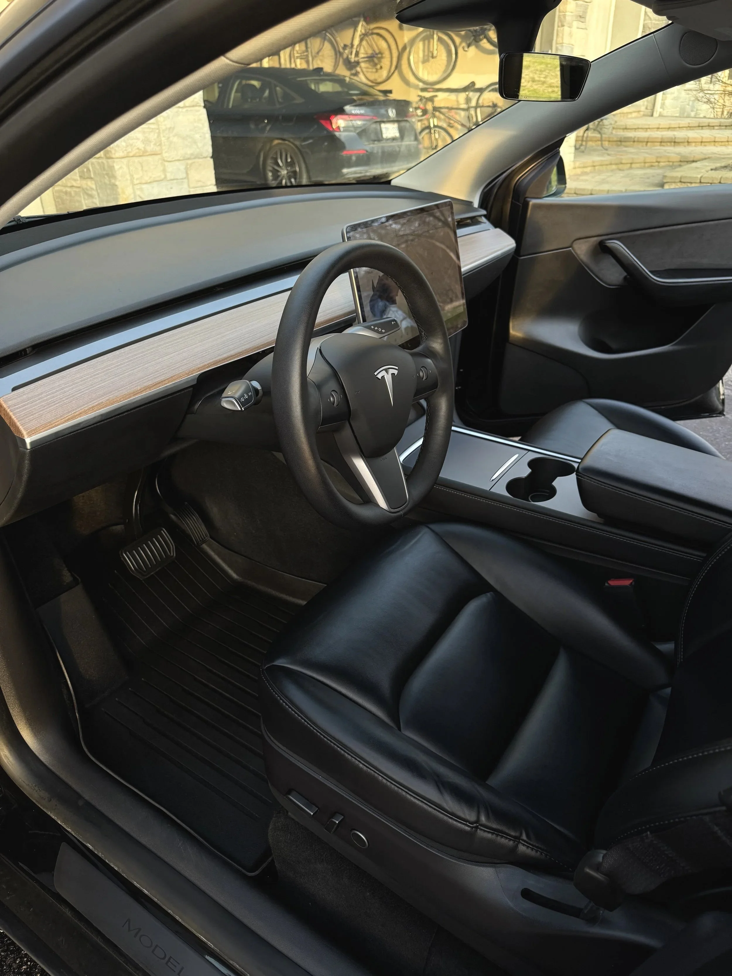 The interior of a Tesla Model 3, featuring a black leather seat, a minimalist dashboard with a large central touchscreen, and a steering wheel with the Tesla logo.