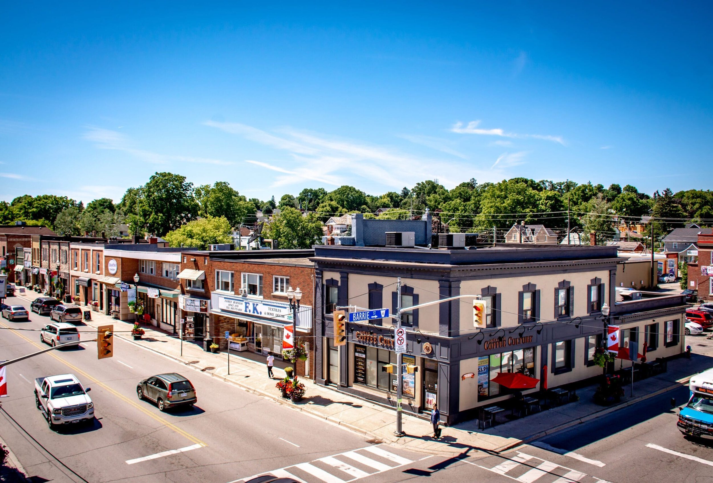A street view in a small town featuring brick buildings with shops, cars on the road, traffic lights, and trees in the background under a partly cloudy sky, with Canadian flags displayed on some buildings.