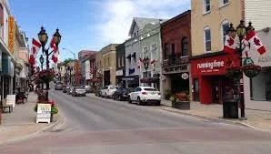A small downtown street with shops and businesses, lined with street lamps and flags, some cars parked along the road, and a few pedestrians visible.